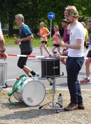 Straßenmusiker mit Schlagzeug auf dem Bürgersteig. Die Straße wurde für einen Marathon abgesperrt. Sechs Joggende joggen an ihm vorbei.
